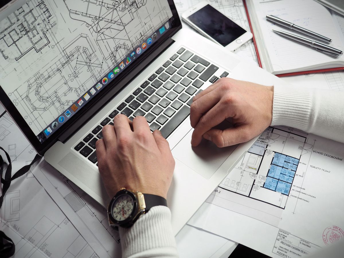 Hands typing on a laptop showing a floor plan, surrounded by rolled architectural blueprints